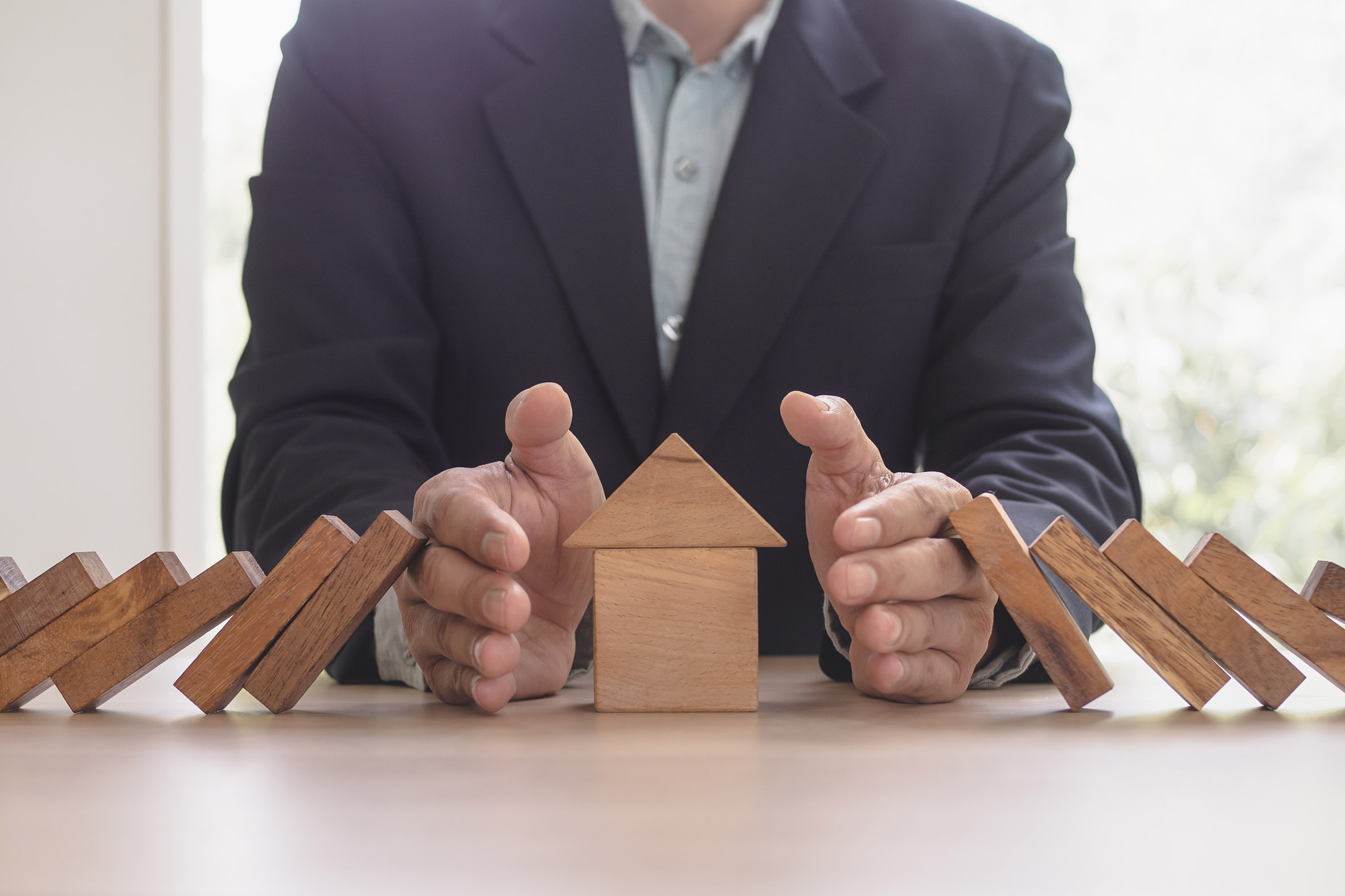 Mann schützt mit seinen Händen ein aus Bauklötzen gebautes Haus vor umfallenden Dominosteinen.